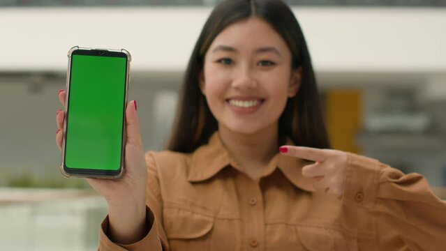 Asian chinese woman hold smartphone in front show mobile phone with green screen mockup advertisement digital app advertise cellphone web service korean girl point finger at empty chroma key display