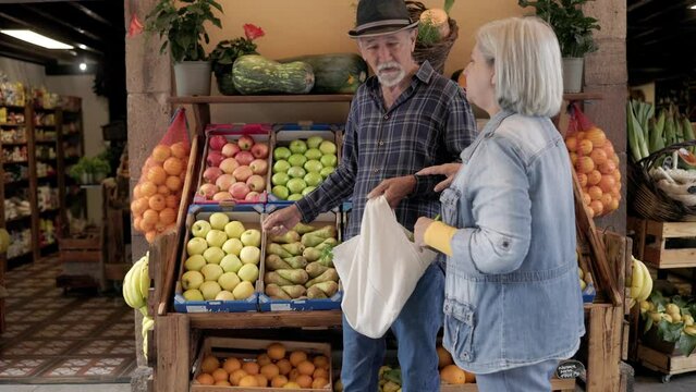 Happy senior couple holding reusable bag of fresh vegetables at local market store - Zero waste food retail