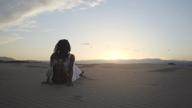 African woman sitting in desert dunes summer day. Girl relaxing on sand. People enjoying sunset in nature. Lady with her back to camera contemplating coastal scenery. Vacations and tourism. 