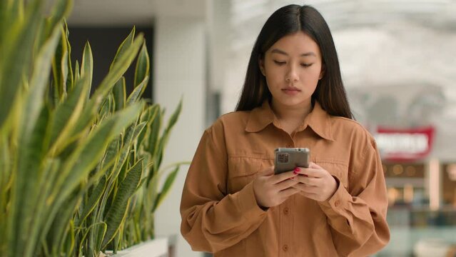 Asian focused woman walking in shopping mall with smartphone look at mobile phone screen Chinese korean girl businesswoman customer walk chatting message online use cellphone app buy in internet store