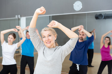 Positive mature blonde doing aerobics exercises in group of adult people, training in fitness center