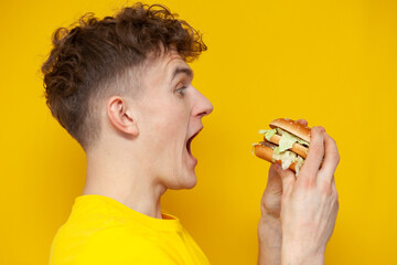 curly guy holding a burger with his mouth open on a yellow background, a man eats fast food,...