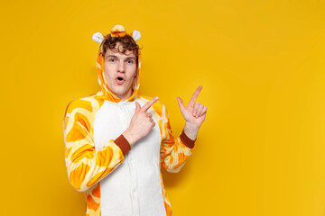 young shocked guy in funny baby giraffe pajamas shows hands on copy space on yellow background