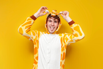 young joyful guy in funny baby giraffe pajamas shows tongue and holds ears on yellow background