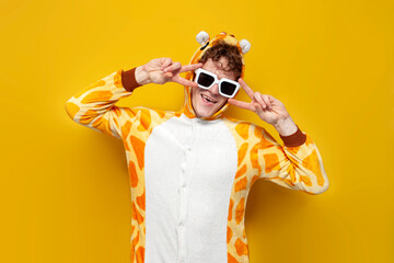 young joyful guy in funny baby giraffe pajamas and glasses is dancing on yellow background