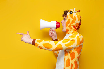 young joyful guy in funny children's giraffe pajamas speaks into megaphone and points his hand to...