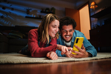 Couple watching funny videos together on a cellphone app lying on the floor at home. Young caucasian wife and husband on the carpet browsing on internet using a smart phone at late night. High quality