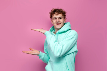 young curly guy in mint hoodie holds copy space with his hands on pink isolated background, man in blue sweatshirt