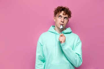 young curly pensive guy in mint hoodie licks spoon and dreams on pink isolated background, hungry...