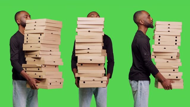 African american man holding pile of pizza boxes stack over greenscreen backdrop, carrying fast food takeaway order in studio. Male model working as deliveryman to give takeout food.