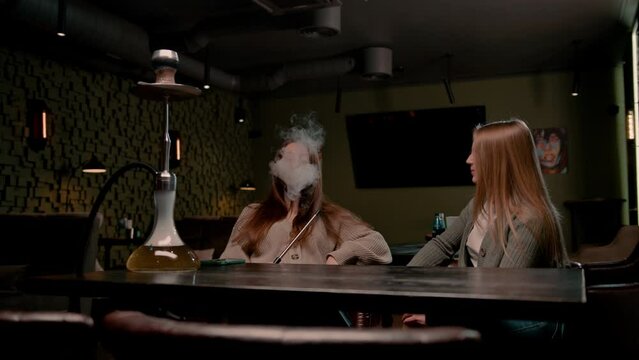 two young female friends are sitting relaxed in a hookah bar smoking hookah and having fun chatting recreation concept