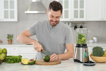 Happy man cutting avocado for delicious smoothie at white marble table in kitchen