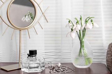 Beautiful snowdrops in vase, perfume, accessories and mirror on wooden table indoors