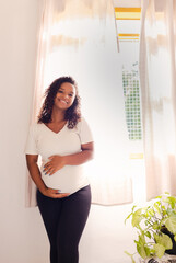 Portrait of a smiling pregnant black Brazilian woman standing indoors with both hands on her belly.