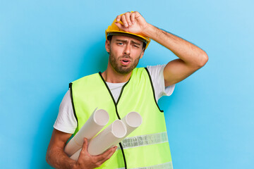 Young tired worker in yellow hardhat holding rolls of paper in his hand, stands isolated over blue backdrop, professional people concept, copy space, high quality photo