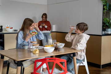 Fototapeta premium Happy mother and son eating together in morning. Teen girl using smartphone while standing away of family having breakfast without her. Cell phone addiction and family relationships