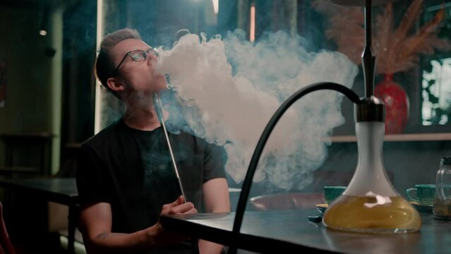 A man in glasses smokes a traditional hookah pipe A man exhales thick smoke in a hookah cafe or lounge bar