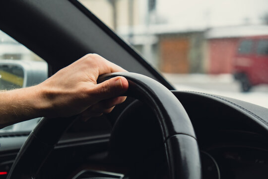 Selective Focus Man's Hand On Steering Wheel, Driving Car In Winter Background. Black Luxury Modern Car Interior. Steering Wheel, Shift Lever And Dashboard. Black Leather Detail Automatic Gear Stick