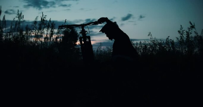 Mountain Bike Rider Silhouette Fixing Electric MTB Bicycle in Meadow Forest. Young Man Repairing Cyclist in Dark Tall Grass Scenic Summer Sunset Dusk. Cinematic Travel Exploration Adventure Sport 4k
