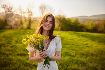 Naklejka na ściany i meble Beautiful woman in a white dress with a bouquet of yellow wildflowers walking in a field 