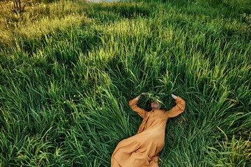 a sweet, calm woman in an orange dress lies in a green field, covering her face with long leaves of grass, enjoying silence and peace. Horizontal photo taken from above
