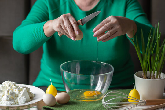 Old woman's hands cracking eggs into glass bowl with knife. Prepare for making cheese pancakes or curd Easter.