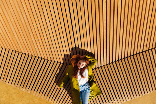 Young red hair woman in coat lying on yellow wooden bench and enjoying sun. Dreaming girl with clothet eyes. Copy space