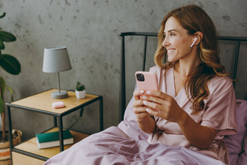 Young woman wears t-shirt pajama earphones sit on bed listen music use mobile cell phone rest relax spend time in bedroom lounge home in own room hotel wake up dream be lost in reverie good mood day.