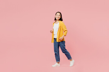 Full body sideways smiling happy young woman of Asian ethnicity wear yellow shirt white t-shirt looking camera walking going strolling isolated on plain pastel light pink background studio portrait.