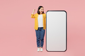 Full body young woman of Asian ethnicity wear yellow shirt white t-shirt big huge blank screen mobile cell phone smartphone with area do winner gesture isolated on plain pastel light pink background.