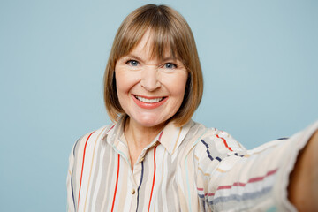 Close up fun elderly woman 50s years old wearing light striped shirt doing selfie shot pov on mobile cell phone isolated on plain pastel blue cyan color background studio portrait. Lifestyle concept.