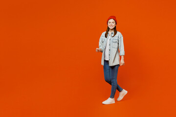 Sideways young smiling fun cool student woman wearing denim shirt white t-shirt red hat looking camera walking going strolling isolated on plain orange background studio portrait. Lifestyle concept.