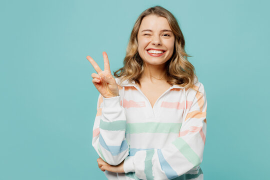 Young smiling cherful fun caucasian blonde woman wearing hoody showing victory sign wink blink eye look camera isolated on plain pastel light blue cyan background studio portrait. Lifestyle concept.