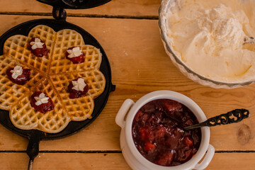 An antique waffle iron with a freshly baked waffle in it decorated with raspberry jam and cream next to it there is a bowl with jam and one with cream