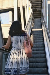 The back view of an Asian woman in white and blue dress is going up an escalator to a sky train station in the morning.