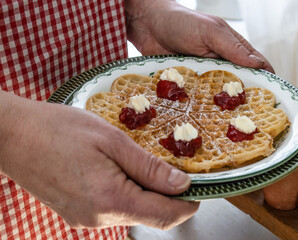 A lady in a red checkered apron holds a plate with a waffle garnished with strawberry jam and cream