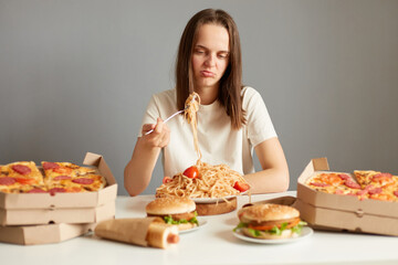Indoor shot of frustrated sad woman with brown hair wearing white T-shirt sitting at table isolated...