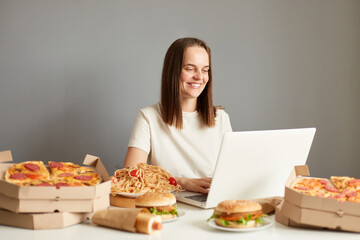 Indoor shot of smiling woman working on laptop computer, typing, communicating online, sitting at table surrounded with fast food isolated over gray background.