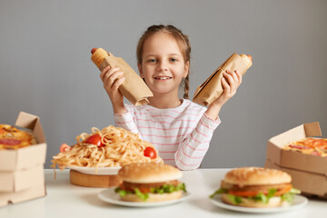 Image of satisfied smiling cute little girl with pigtails sitting at table isolated over gray background, holding hot dogs, having cheat meal, looking at camera with happy face.