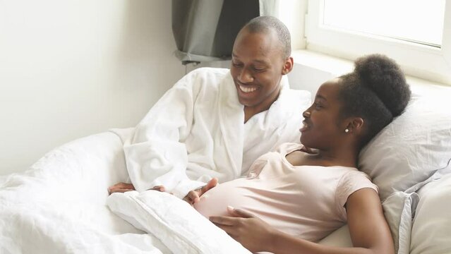 Black man hugs his pregnant wife on bed, and touches her stomach. Happy relationship, pregnancy concept.