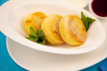 Round cottage cheese pancakes in a white plate on a blue background. Food breakfast. Healthy food. Sour cream sauce and mint. Top view closeup. Dessert meal. Curd cheese pancake ricotta fritters.