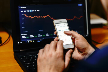 Businessman trader investor analyst using mobile phone app analytics for financial market analysis,phone with graphs and diagrams on screen,using smartphone.sitting at work desk with laptop.