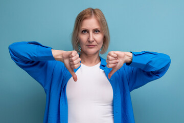 portrait of serious blonde 50s woman in blue stylish shirt showing dislike on studio background