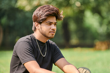 Attractive young indian man portrait in black t shirt and silver neck chain sitting on green lawn...