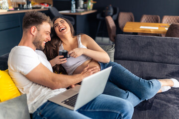 Fototapeta premium Expecting new parents are sitting on the couch planning the decor for the new children's room, the laptop is on his lap while he is touching her belly,she holds a cup of tea in her hand