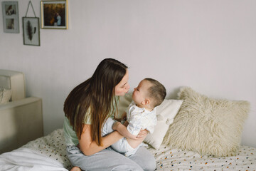 Home portrait of a baby boy with mother on the bed relaxing and playing in the bed at the weekend together, lazy morning, warm and cozy scene. Mom holding and kissing her child. Mother day concept