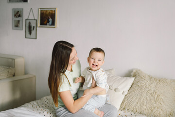 Home portrait of a baby boy with mother on the bed relaxing and playing in the bed at the weekend together, lazy morning, warm and cozy scene. Mom holding and kissing her child. Mother day concept