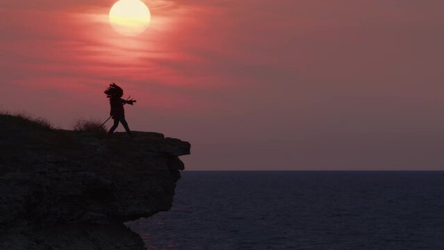 Silhouette of Red Indian chief in headdress with feathers on edge of cliff on shore of ocean or sea calls and looks into distance on background of amazing sunset sky with sun.