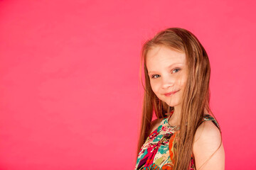 Face of smiling little girl in colorful t-shirt posing at pink background, looking at camera. Portrait of young lady with long hair expressing emotion. Childhood emotional concept. Copy ad text space