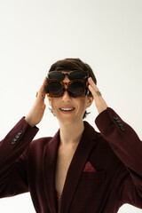 cheerful woman in burgundy brown blazer fitting on several stylish sunglasses isolated on grey.